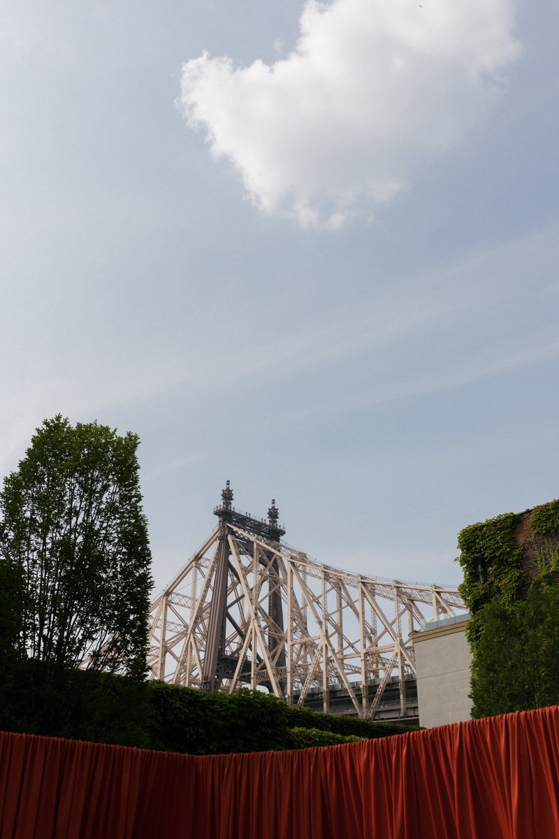 A wide shot captures a section of a large metal bridge, likely the Queensboro Bridge, partially obscured by lush green t