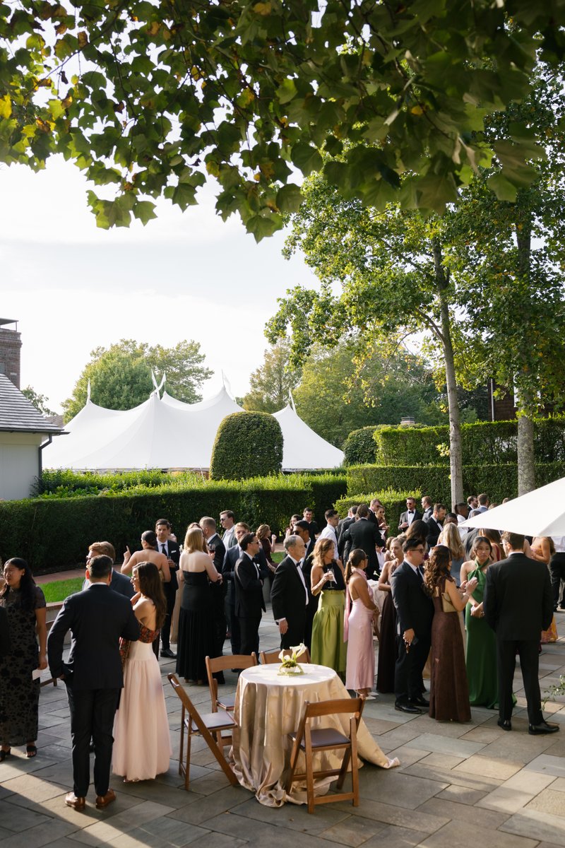 Guests in formal attire mingle on an outdoor patio during a cocktail reception. A large white tent and lush green landsc