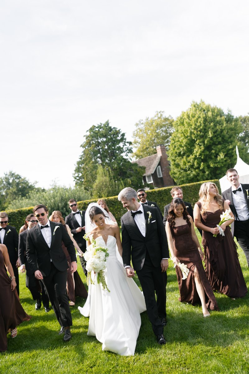 A joyful wedding party, led by the smiling bride and groom holding hands, walks across a lush green lawn. Bridesmaids in