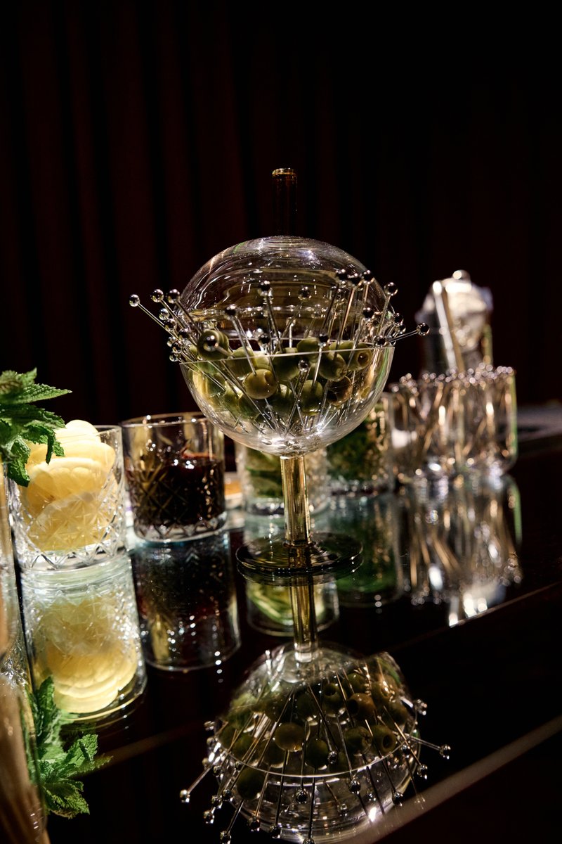 A close-up shot of an elegant olive dispenser, filled with green olives on decorative picks, standing on a reflective da