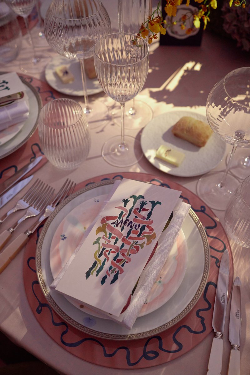 An elegantly set table features a decorative menu card placed on a layered plate setting, surrounded by crystal glasswar