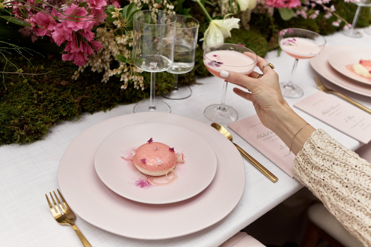 A beautifully arranged table setting features a delicate pink dessert and a matching cocktail, with a hand reaching for 