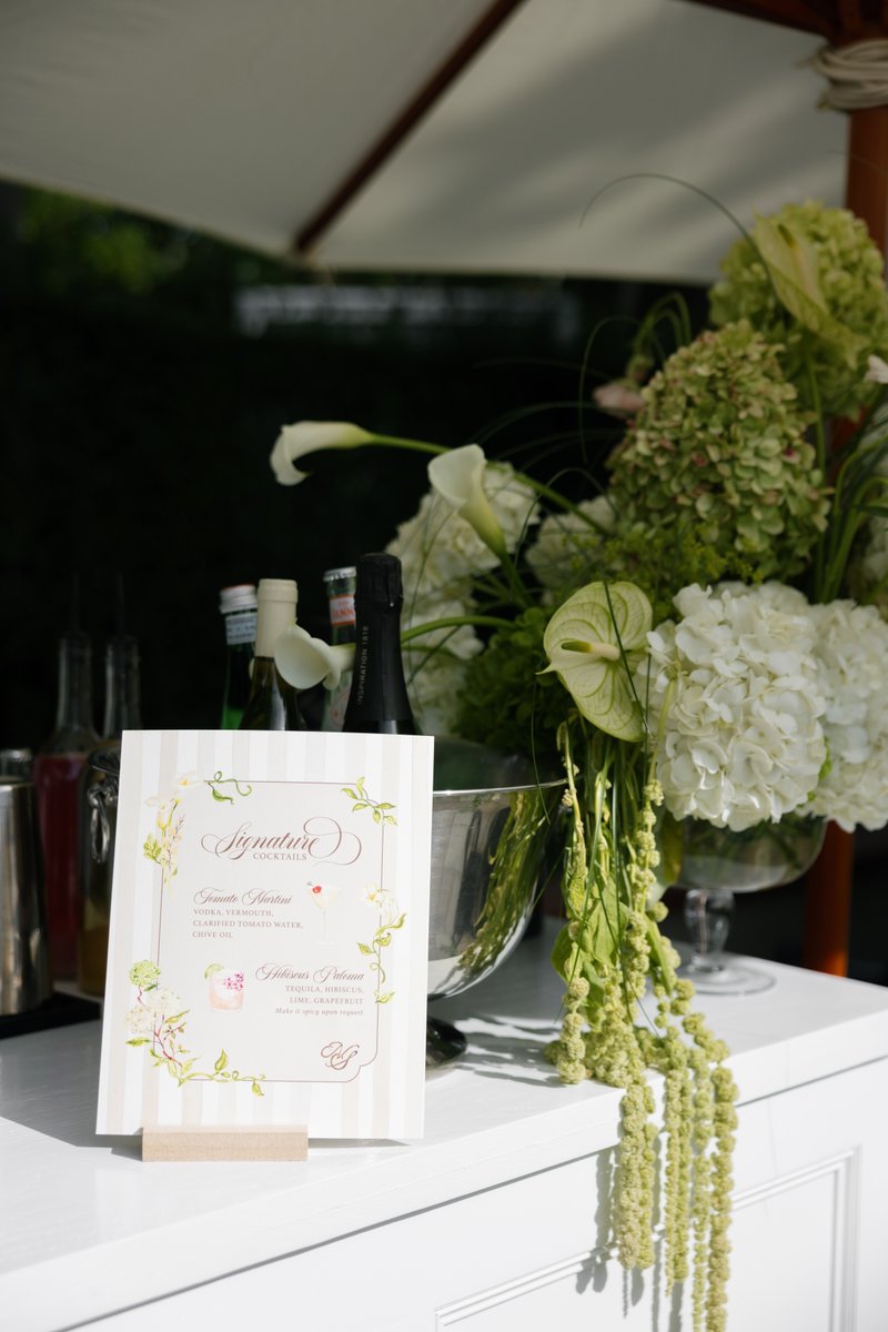 A beautifully arranged bar station features a 'Signature Cocktails' menu card, bottles, and an elegant floral arrangemen