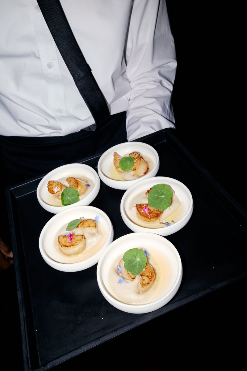 A server, partially visible in a white shirt and black apron, holds a black tray presenting five small, elegantly plated