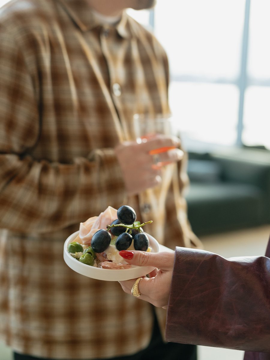 A person holds a small plate of appetizers featuring dark grapes, green olives, and sliced charcuterie, while another in