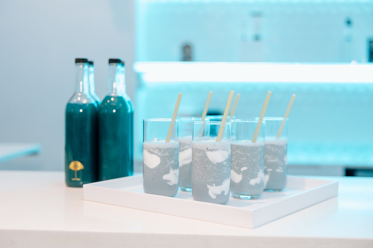A collection of blue-grey smoothies with white swirls in clear glasses, each with a straw, are neatly arranged on a whit