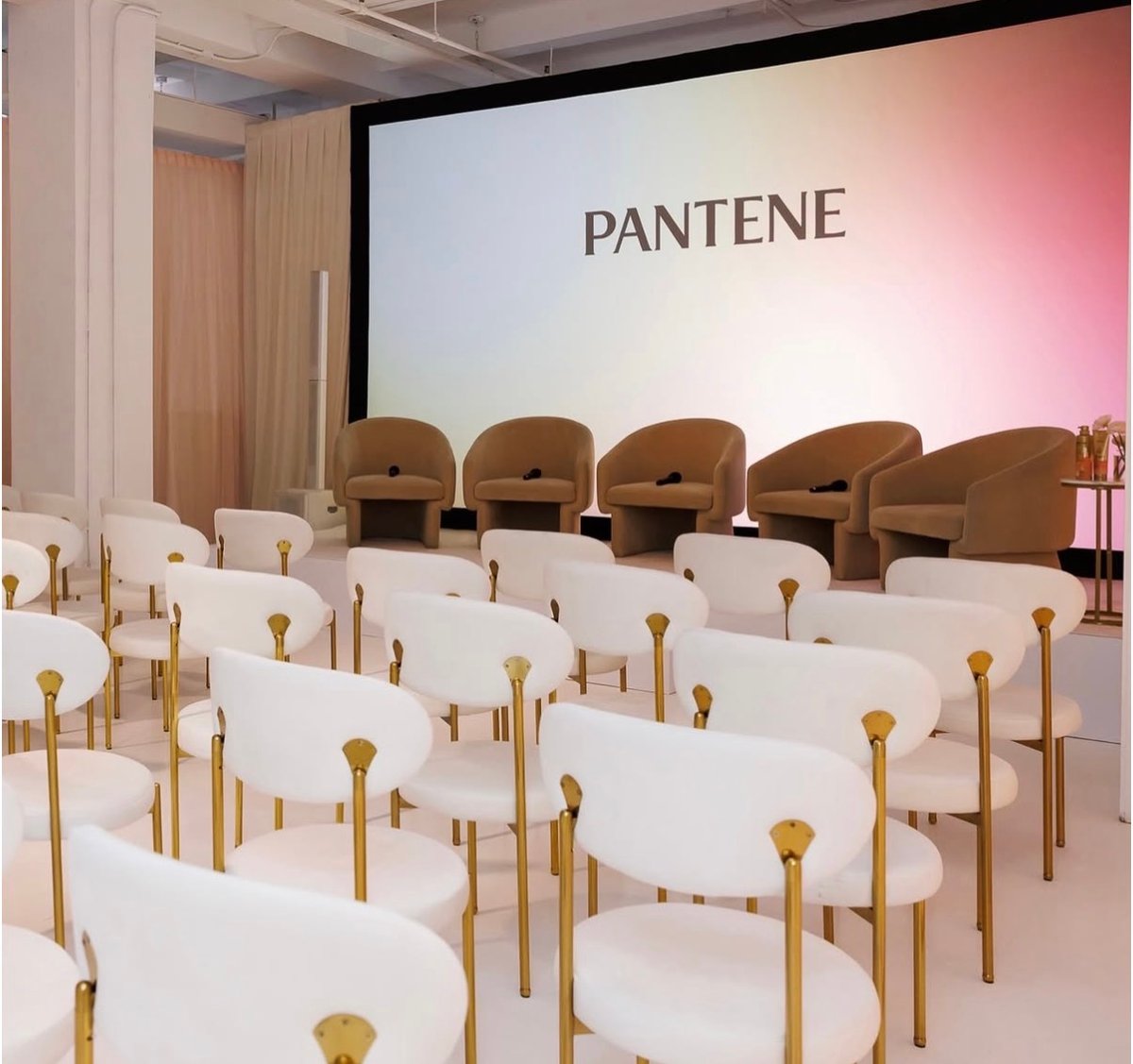 A modern corporate event space is set up for a presentation or panel discussion. The large screen displays the 'PANTENE'