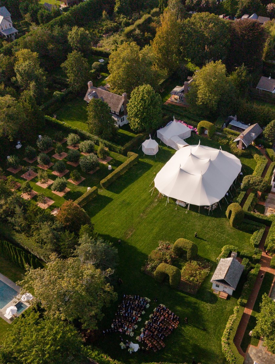 An expansive aerial view captures a large outdoor wedding event set on a lush green property. A grand white tent dominat