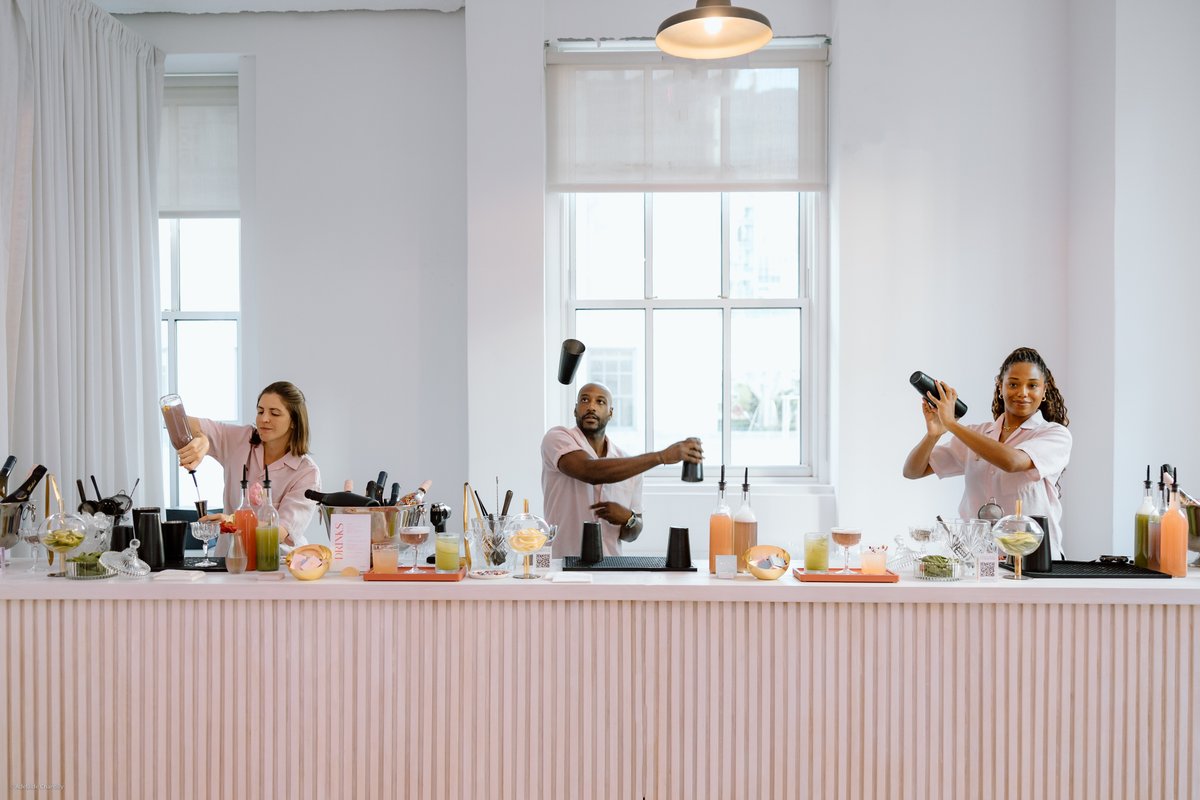Three bartenders are actively preparing a variety of cocktails behind a modern, light pink fluted bar in a bright, minim