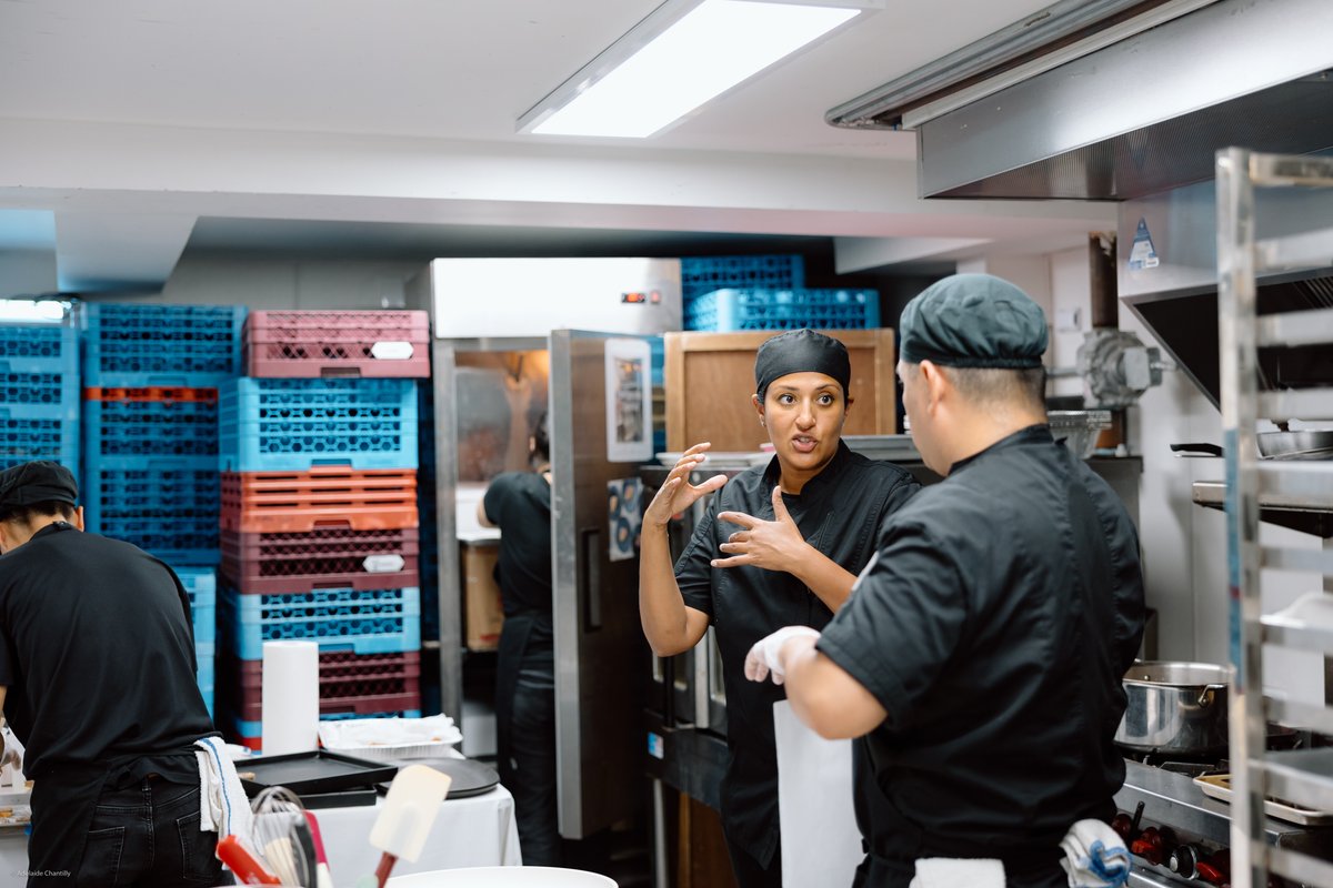 Three chefs are busy in a commercial kitchen. In the foreground, a female chef in a black uniform and head covering is a