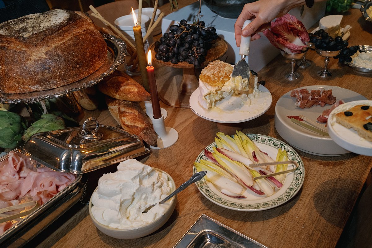 A close-up shot captures a lavish grazing table laden with artisanal foods. A hand is actively serving a generous portio