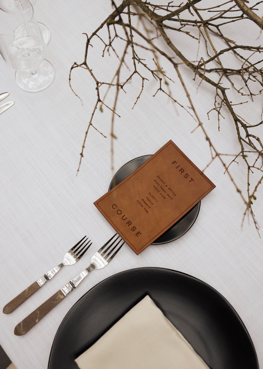 An elegant overhead shot captures a minimalist table setting with a brown leather menu card resting on a black plate, ac