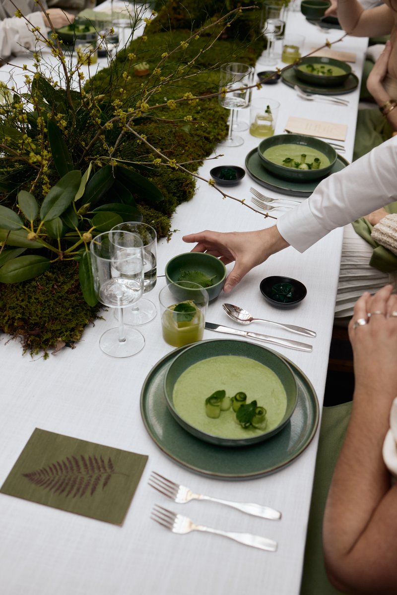 A close-up shot captures an elegantly set dining table adorned with a lush green moss and branch runner. A server's hand