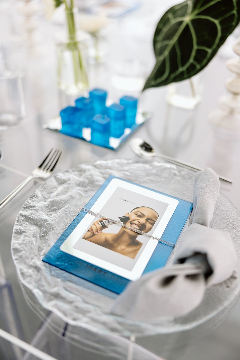 A modern table setting features a clear glass charger plate holding a blue promotional box with a card. Cutlery and a li