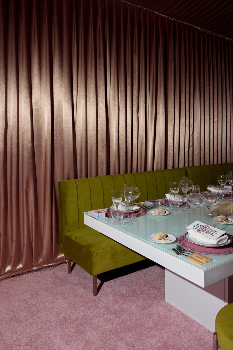 A vibrant dining area is set with a long white table, green velvet banquette seating, and individual place settings. The