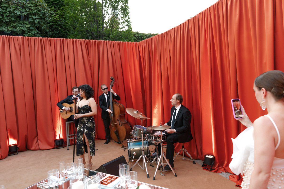A band performs on an outdoor stage with vibrant burnt orange drapes as a backdrop, while a guest in the foreground capt