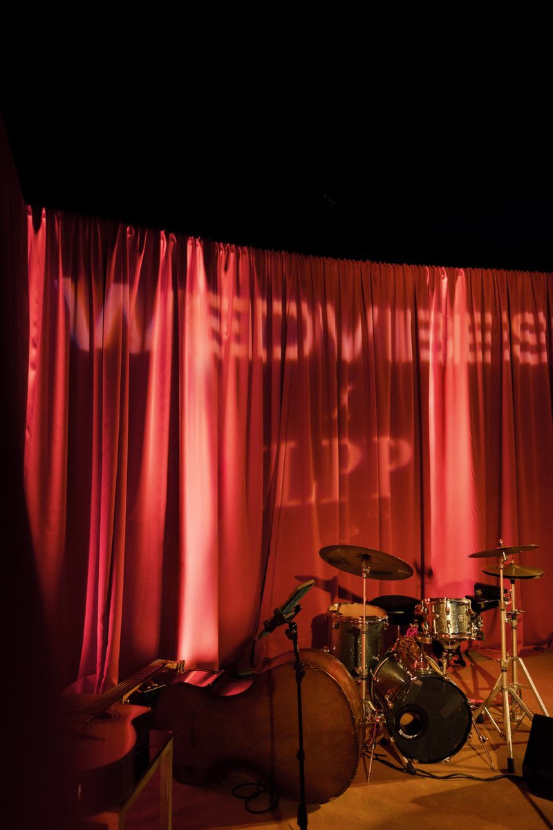 A stage is set with musical instruments, including a drum kit, an upright bass, and a guitar, against a backdrop of vibr
