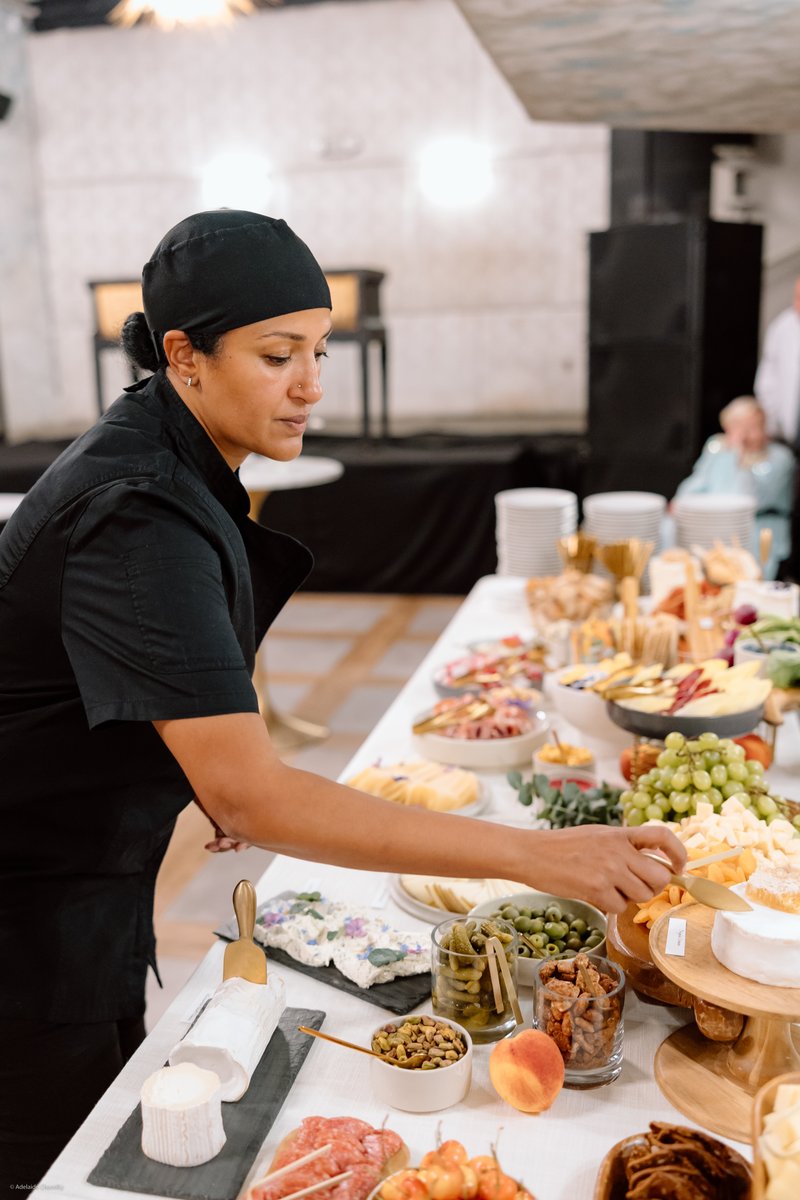 A female chef, dressed in a black uniform and head covering, stands holding a microphone and speaking. She is positioned