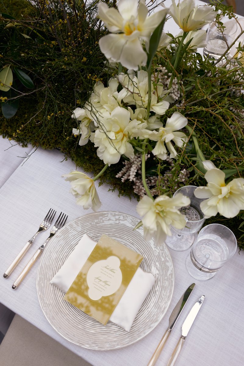 A beautifully arranged table setting features a delicate white and cream floral centerpiece with lush green foliage, cre