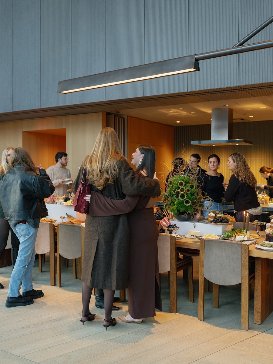Guests are gathered around a long wooden table laden with food, engaging in conversation. The setting appears to be a mo