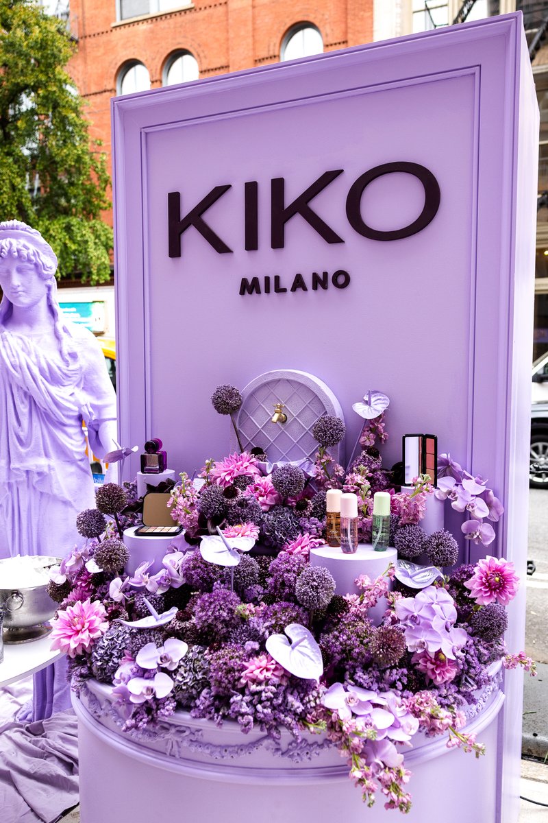 A lavish KIKO Milano product display is set up outdoors, featuring various cosmetic items nestled among a vibrant arrang
