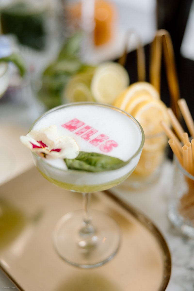 A vibrant cocktail, likely a sour, is presented in a coupe glass with a frothy white top, adorned with a delicate flower