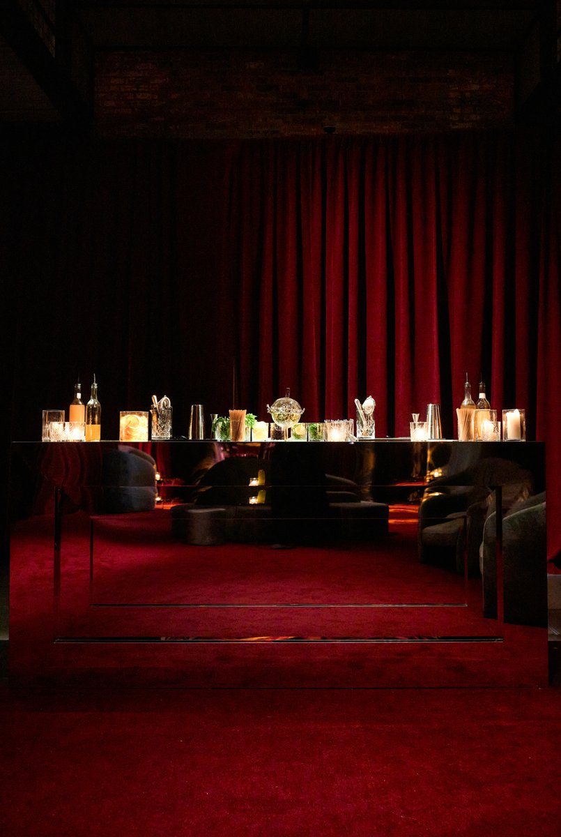 A sophisticated, mirrored bar station is set against a dramatic deep red velvet curtain backdrop in a dimly lit venue. T