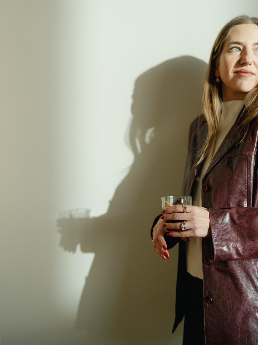 A woman in a maroon leather jacket holds a cocktail in a rocks glass, looking thoughtfully to the side. A dramatic shado