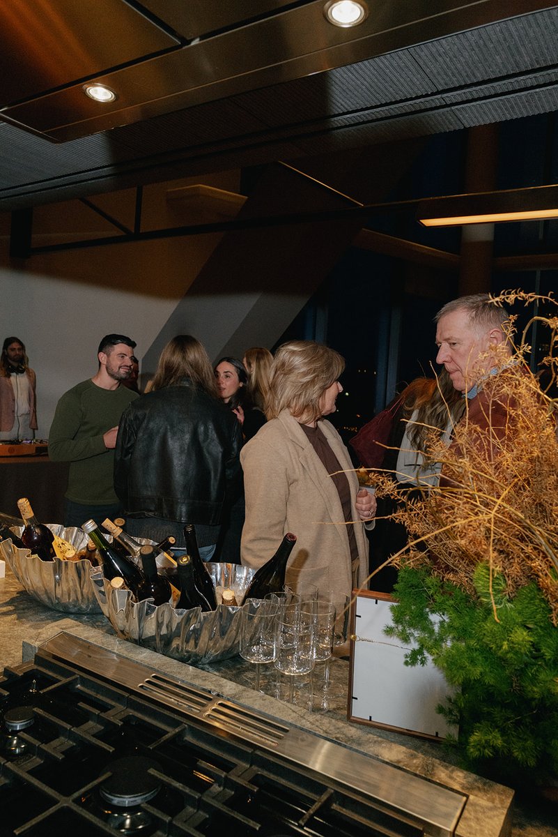 Guests are seen mingling around a wine station set up on a kitchen counter, featuring multiple bottles in ice buckets an