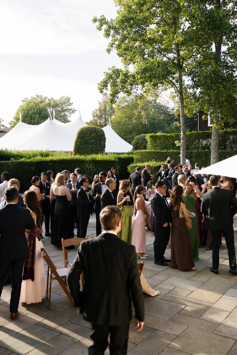A large group of elegantly dressed guests are mingling and socializing outdoors on a paved patio. A prominent white even