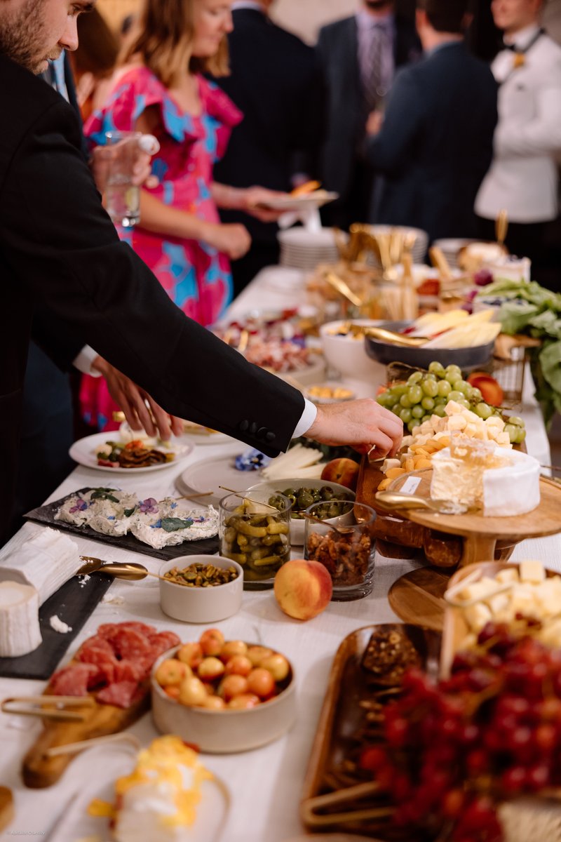 Guests are seen serving themselves from a lavish buffet table laden with an assortment of cheeses, charcuterie, fruits, 