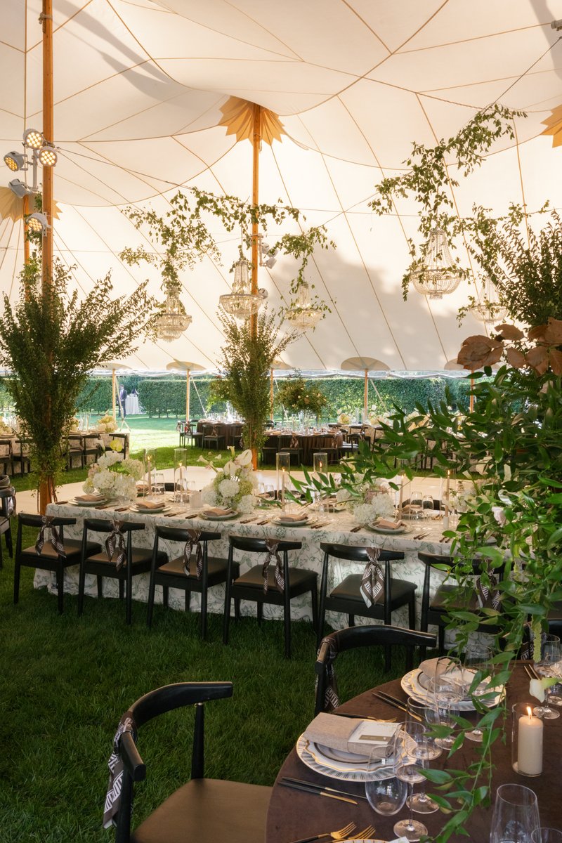 This wide shot captures an elegant event tent set up on a lush green lawn. Multiple tables are meticulously arranged wit
