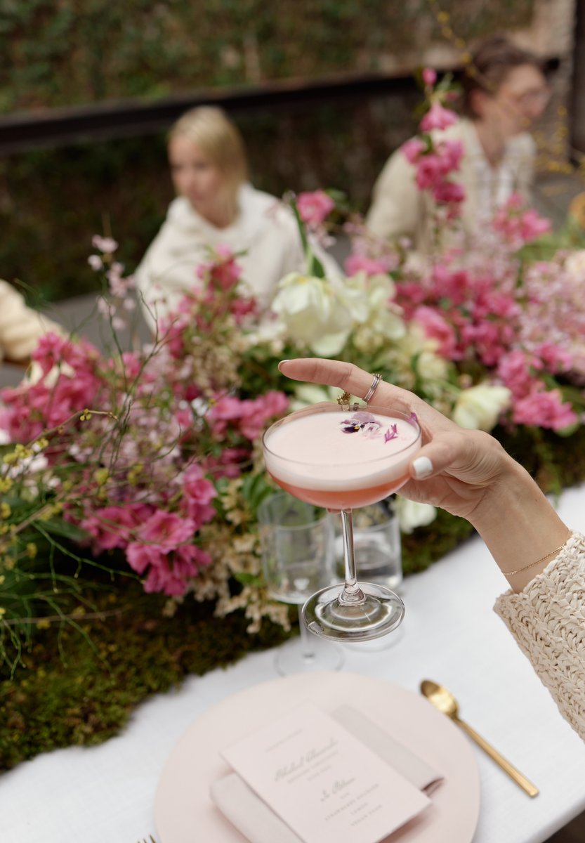 A person's hand, adorned with rings, holds a delicate pink cocktail garnished with small purple flowers, positioned over