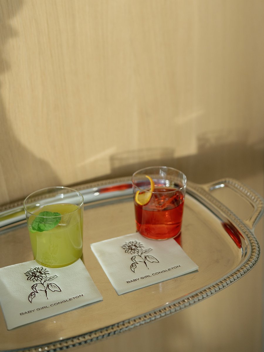 Two distinct cocktails, one green with mint and one red with an orange peel, are presented on a polished silver tray. Ea