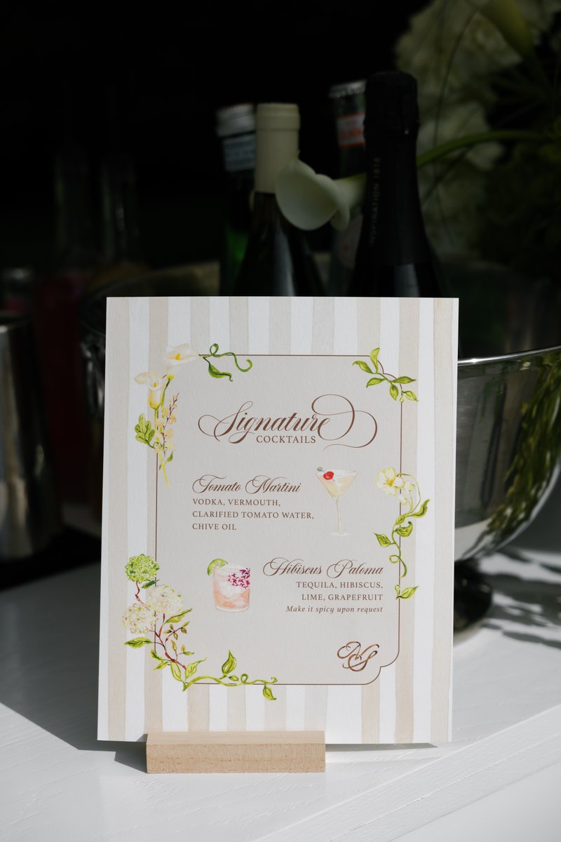 A beautifully designed signature cocktail menu stands on a white table, featuring 'Tomato Martini' and 'Hibiscus Paloma'