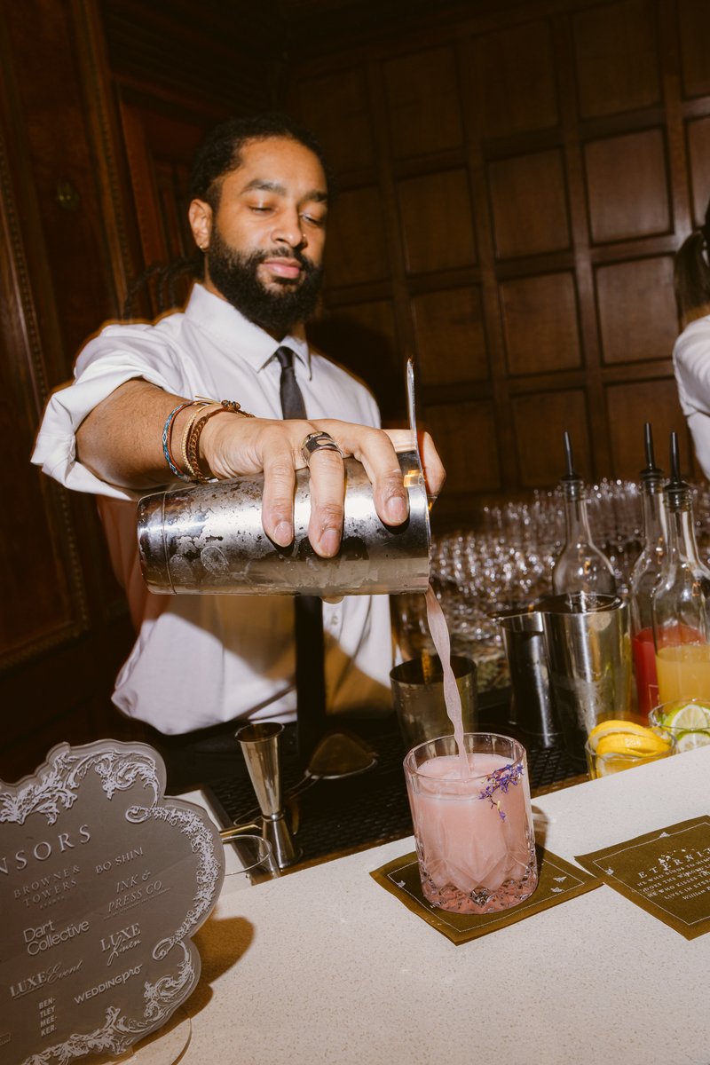A bartender with a beard and dreadlocks, wearing a white shirt and tie, is intently pouring a pink cocktail from a shake