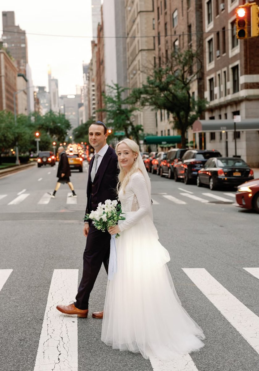 A newlywed couple, the bride in a white gown holding a bouquet and the groom in a dark suit, are walking across a crossw