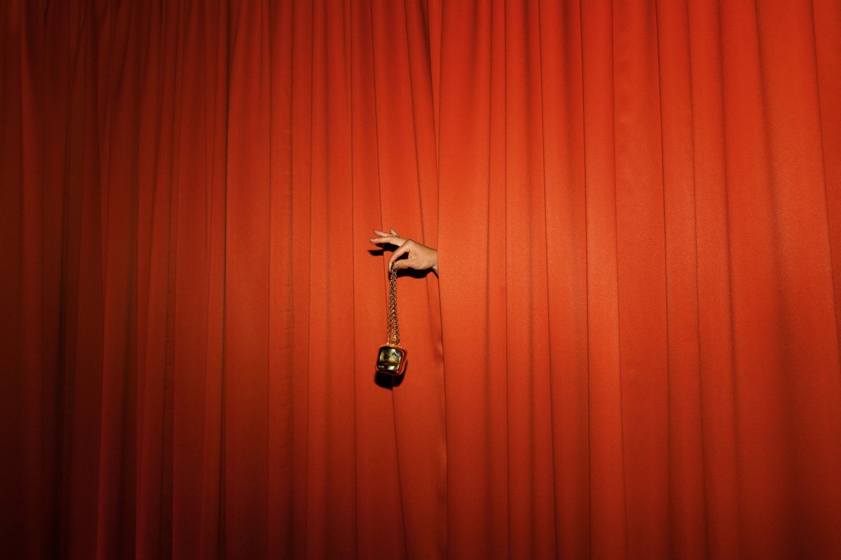 A human hand emerges from vibrant red stage curtains, delicately holding a small, golden, cube-shaped decorative object 