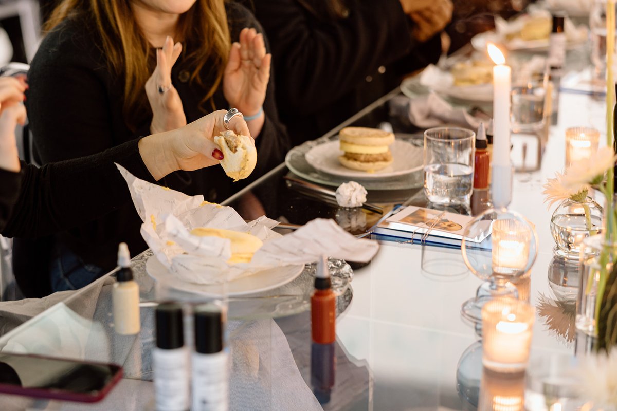 Guests are seated at a long, reflective table, enjoying individual meals. One person in the foreground holds a small san