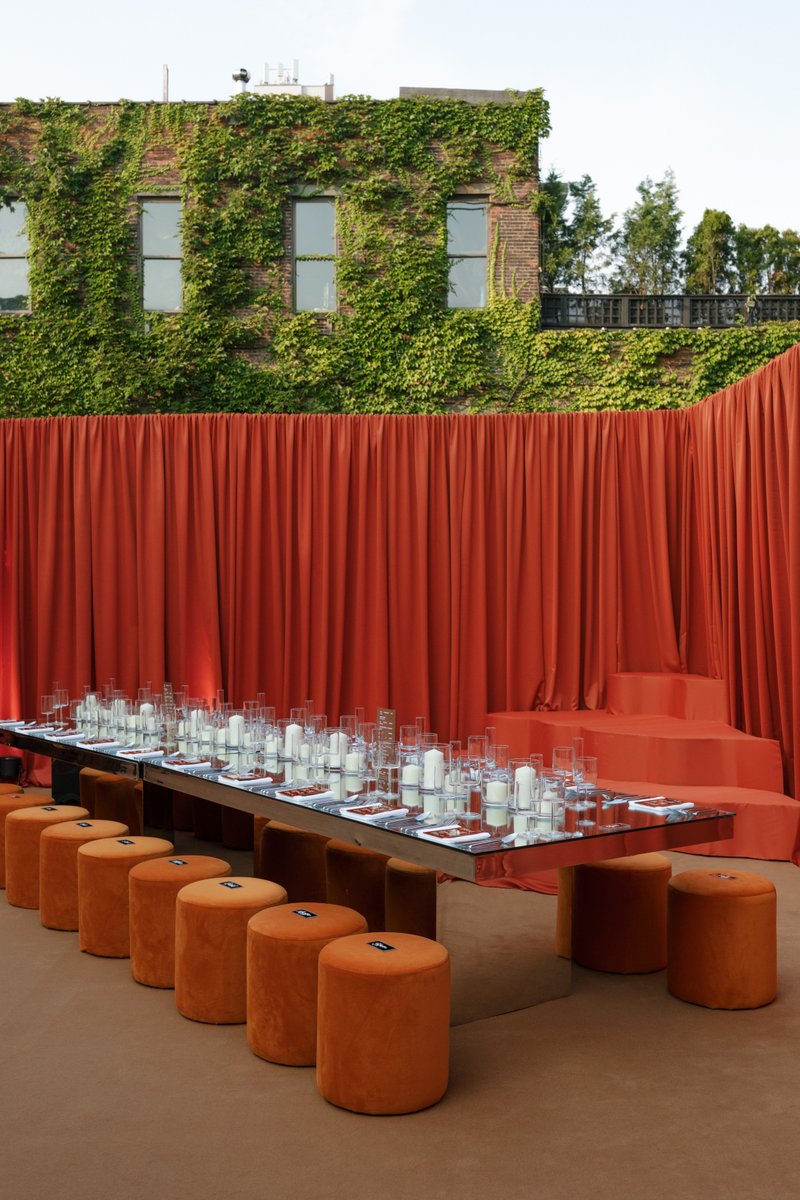 A long, reflective dining table is meticulously set with glassware and candles, flanked by orange velvet stools. The set