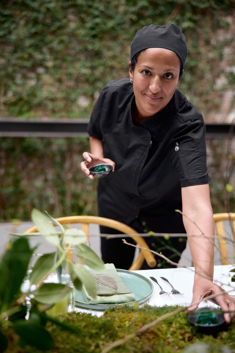 A chef in a black uniform and hat is meticulously arranging a table setting, looking directly at the camera with a sligh