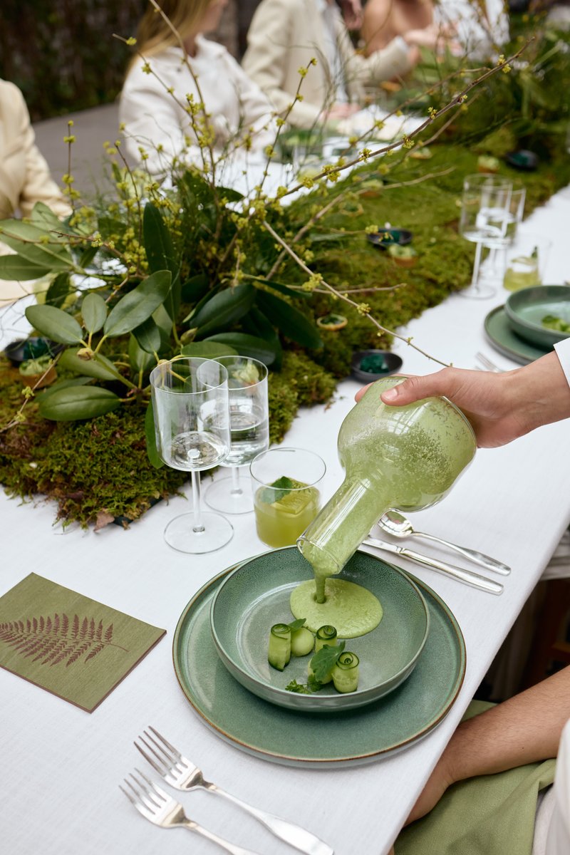 A close-up shot captures a hand pouring a vibrant green soup from a unique glass pitcher into a matching green bowl, gar