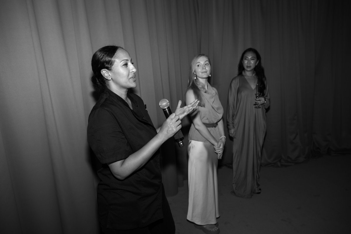 A woman in a dark shirt speaks into a microphone, gesturing with her hand, while two other women stand attentively behin