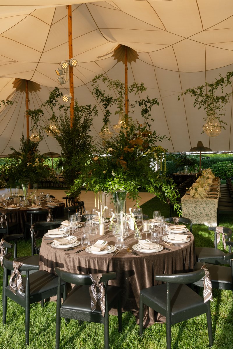 This wide shot captures an elegantly set event space under a large tent, featuring round tables draped in dark brown lin