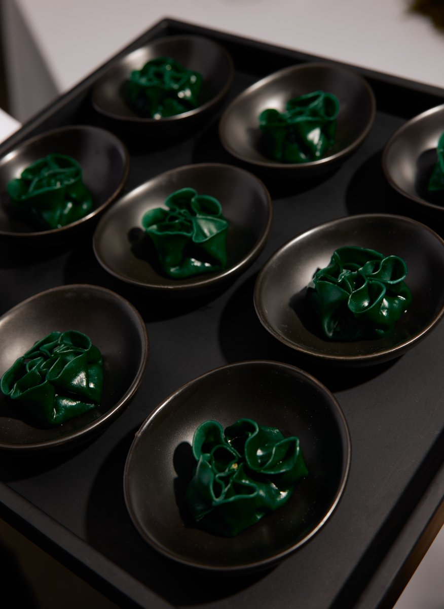 A tray of meticulously arranged dark green appetizers, possibly pasta or dumplings, served in individual small black bow