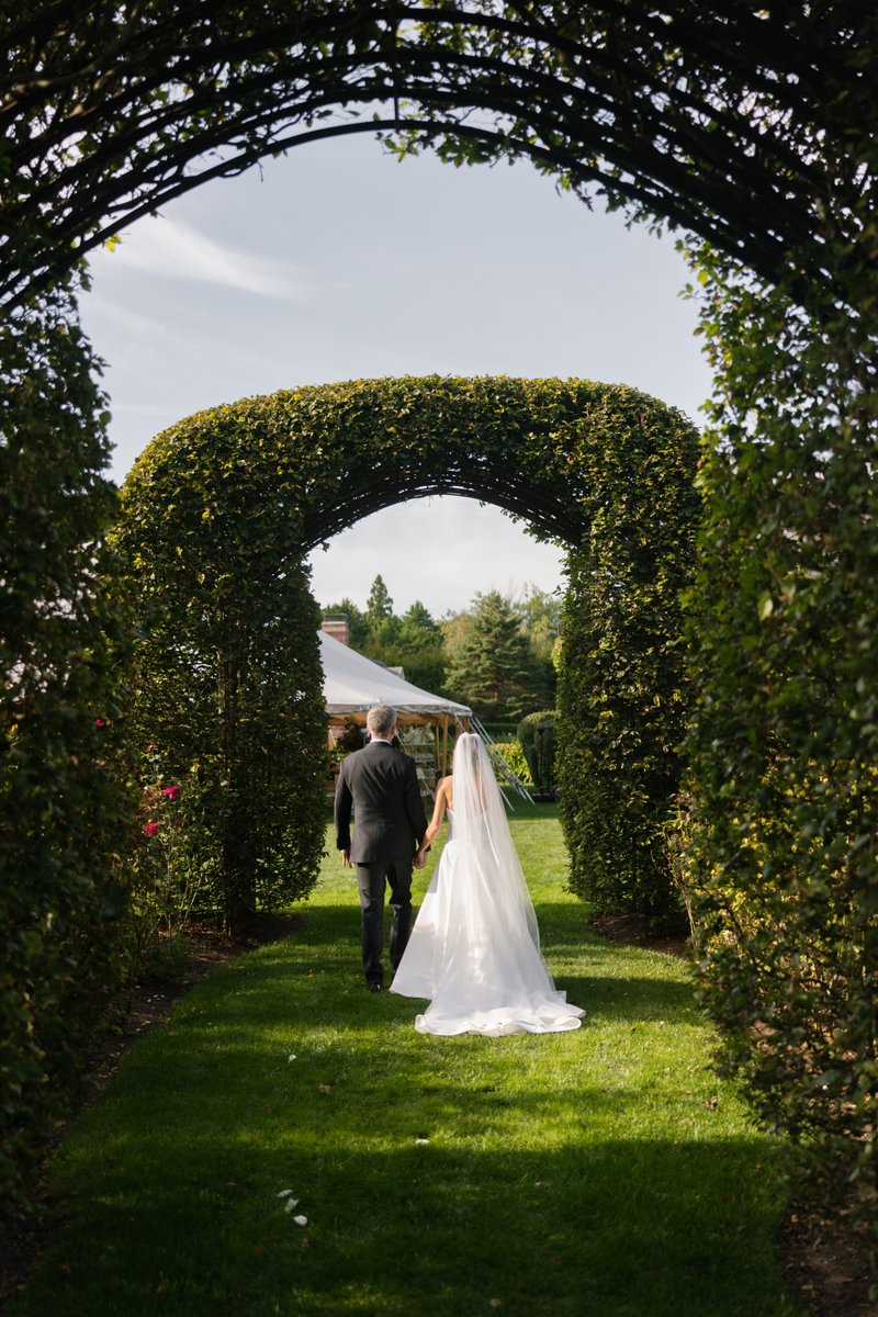 A couple, likely a bride and groom, walks hand-in-hand through a lush green garden archway. A white event tent is visibl