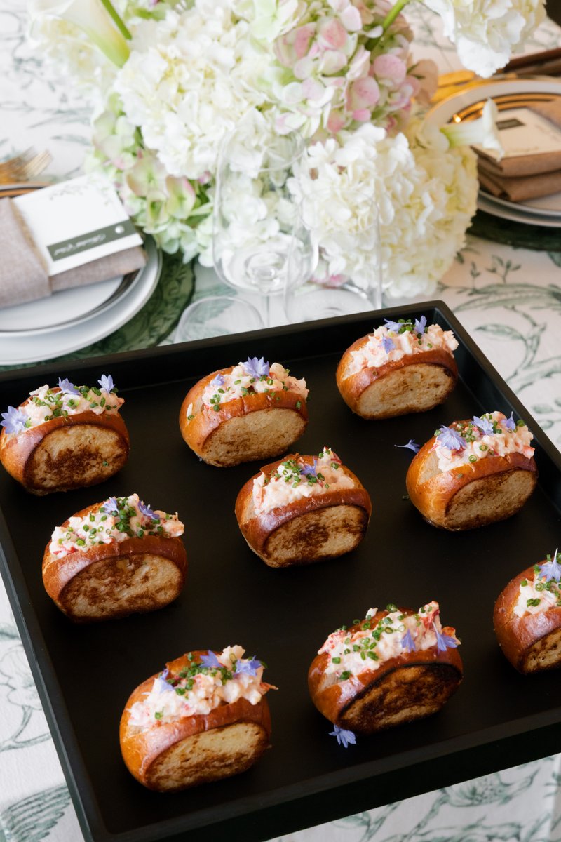 A black tray showcases several mini lobster rolls, beautifully garnished with chives and small purple edible flowers. Th