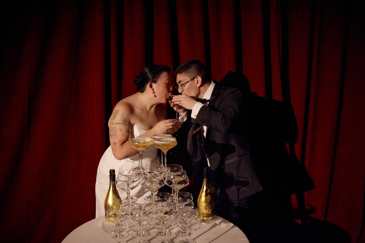A couple, likely a bride and groom, shares an intimate moment drinking from a champagne tower. They are positioned in fr
