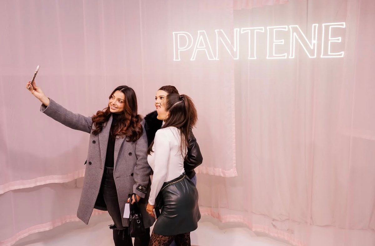 Three women are posing for a selfie in front of a light pink curtain backdrop with a 'PANTENE' neon sign. The woman on t