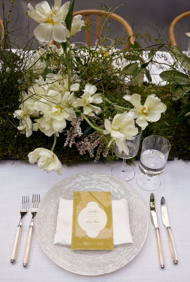 A close-up shot of an elegantly set table featuring a lush, low floral centerpiece with white tulips and various greener